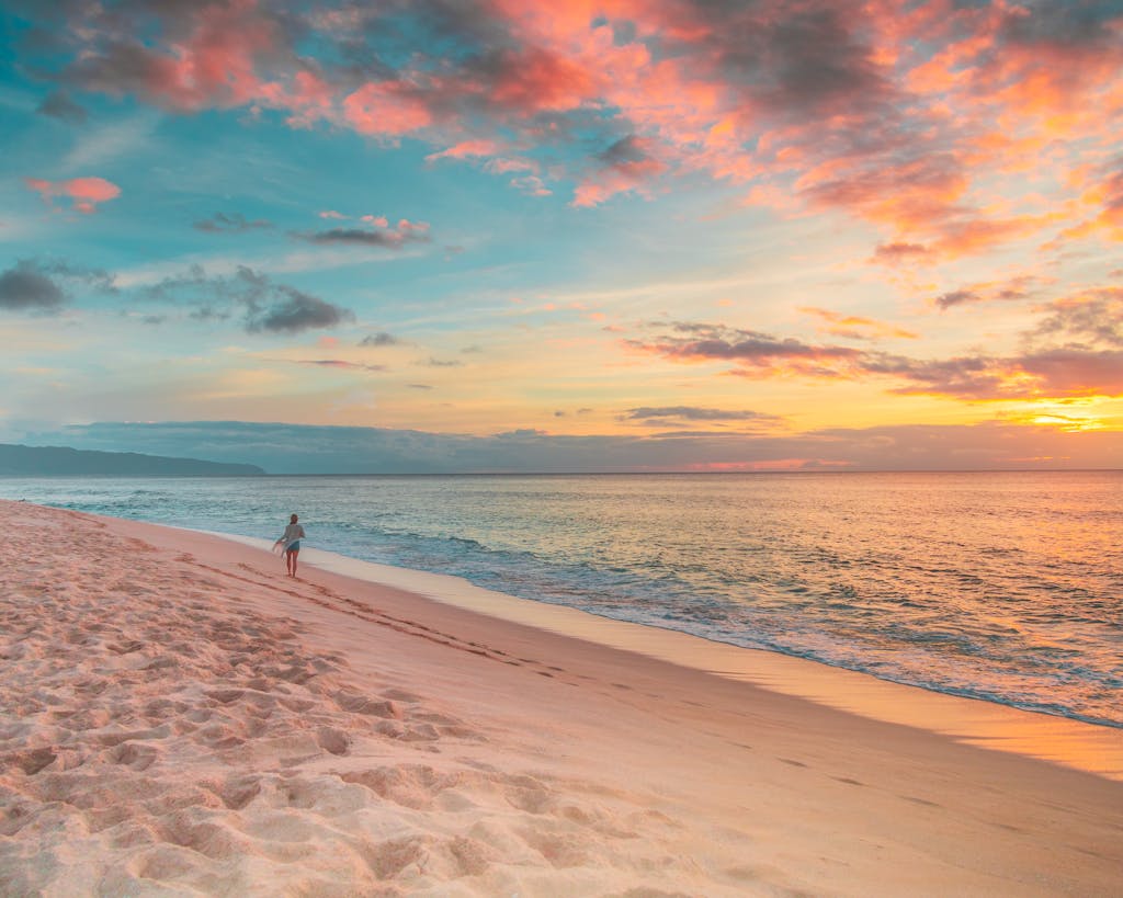 Tranquil sunset view on a Hawaiian beach with a lone person walking along the shoreline.