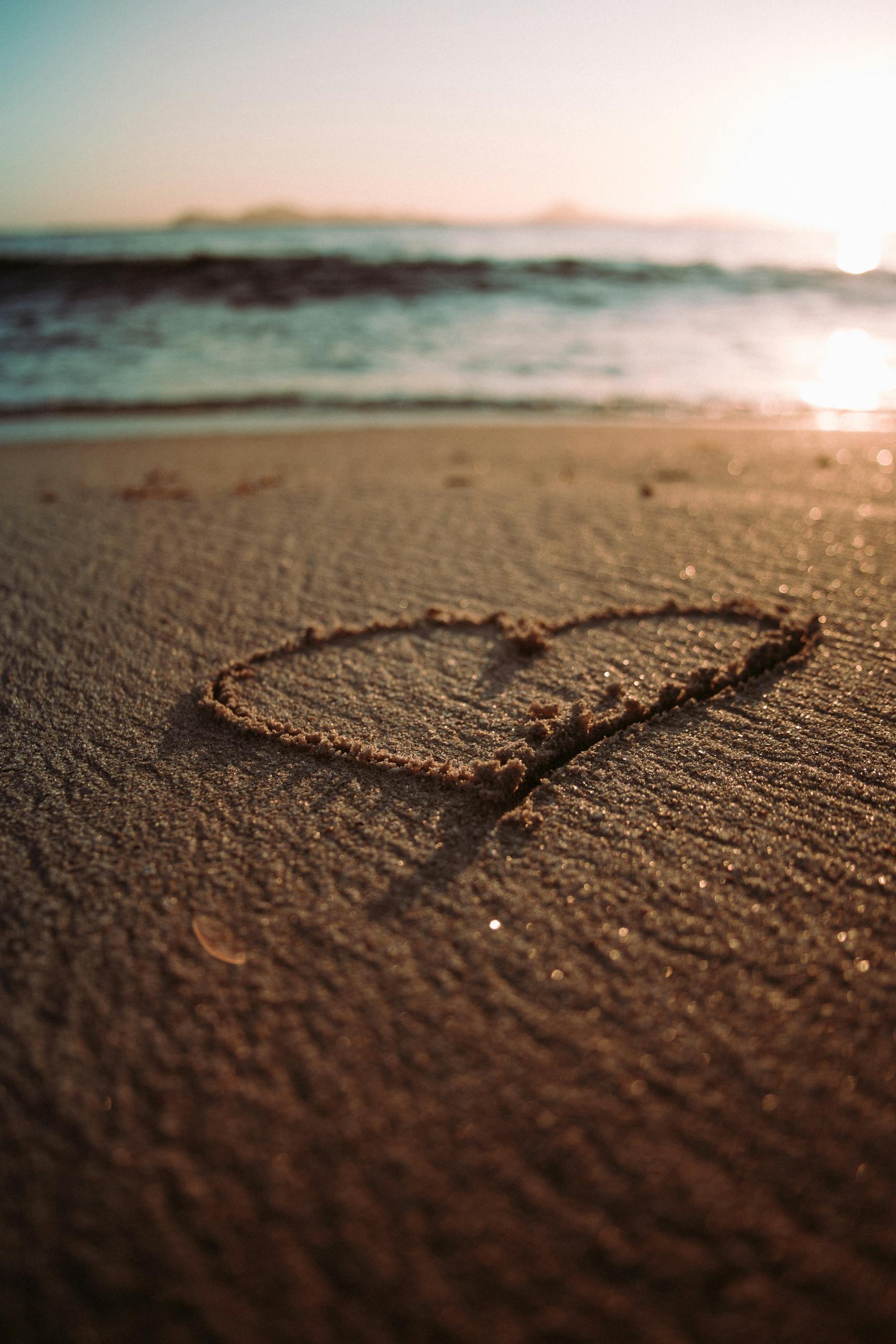 A heart shape drawn in the sand on a beach at sunrise with waves in the background.