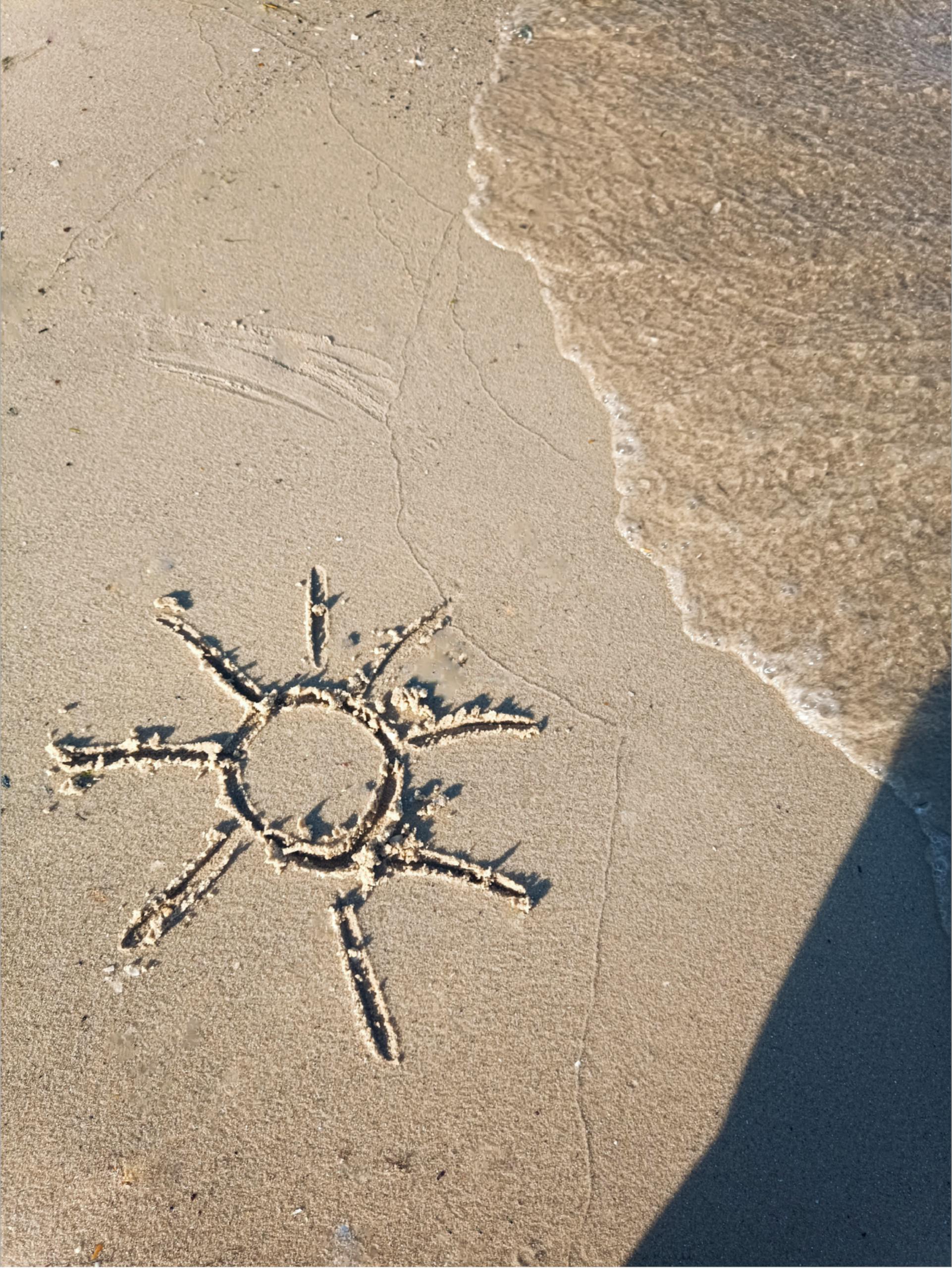 A sun drawing etched in the beach sand near the ocean waves, suggesting a sunny beach day.