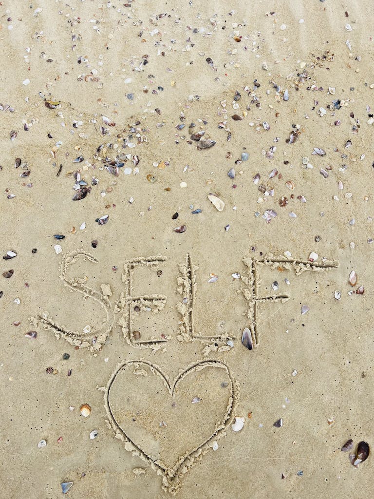 The word 'self' with a heart shape drawn in sand amidst scattered seashells on a beach.