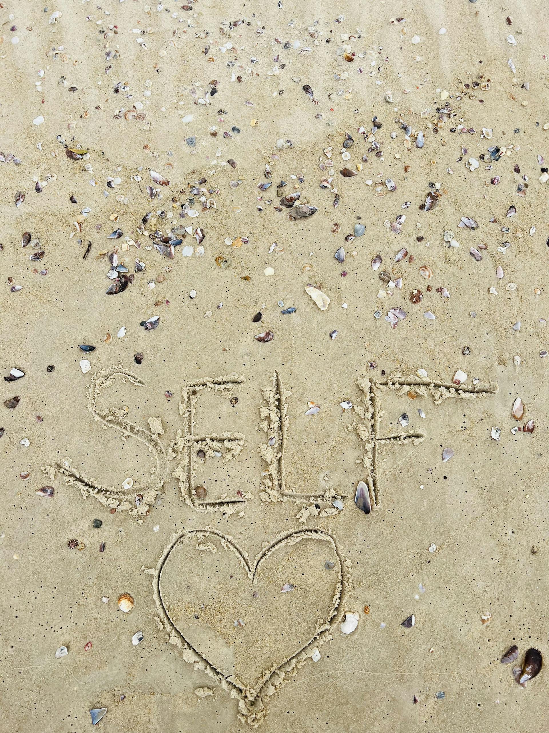 The word 'self' with a heart shape drawn in sand amidst scattered seashells on a beach.